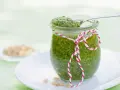 Un pot de pesto vert vif, décoré d'une ficelle rouge et blanche, prêt pour une recette pesto traditionnel. Quelques pignons de pin à côté.