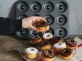 Une main tient un donut glacé au chocolat, prêt à être dégusté. Une belle collection de donuts au four, certains saupoudrés de sucre glace, d'autres de glaçage au chocolat et de vermicelles colorés, attendent sur une grille.