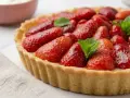 Tarte aux fraises fraîchement préparée, garnie de fruits rouges brillants et de feuilles de menthe, sur un fond clair.
