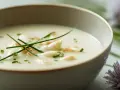 Un bol de velouté d'asperges blanches, garni de brins de ciboulette et de morceaux d'asperges. Des fleurs d'ail ornent le bord.