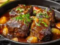 Quatre morceaux de joue de bœuf braisée, tendres et juteux, mijotent dans une sauce riche avec des oignons caramélisés et des herbes fraîches.