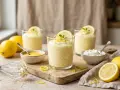 Trois verres de mousse au citron, décorés de rondelles de citron et de menthe. Des citrons frais, un fouet et des petits bols complètent cette scène gourmande.