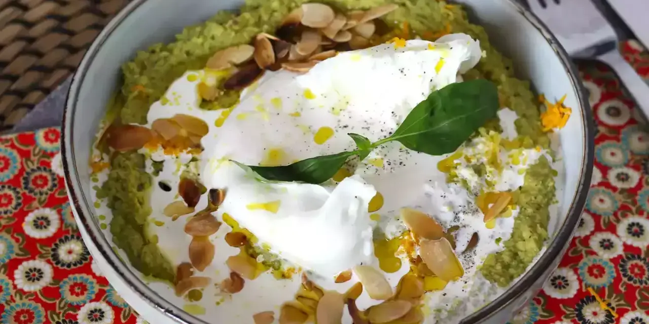 Un bol de purée verte, peut-être une sauce petit pois, garni d'un œuf poché, de crème, d'amandes effilées et d'une feuille de basilic.