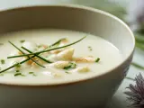 Un bol de velouté d'asperges blanches, garni de brins de ciboulette et de morceaux d'asperges. Des fleurs d'ail ornent le bord.