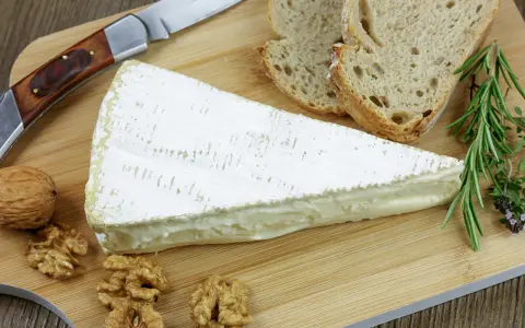 Tranche de fromage à croute lavée crémeux, accompagnée de pain, noix et herbes aromatiques sur une planche en bois.