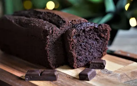 Un délicieux cake au chocolat, moelleux et intense, coupé en tranches sur une planche en bois, avec des carrés de chocolat noir en garniture.