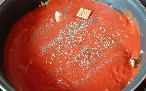 Un coulis de tomates mijote dans une poêle, agrémenté d'herbes et d'un cube de bouillon.