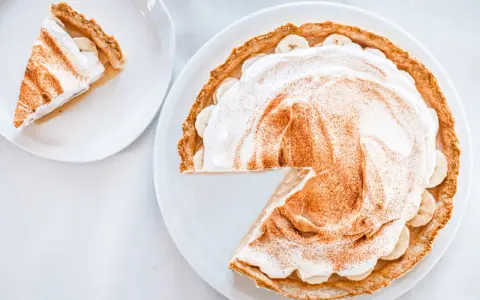 Une part de tarte banoffee traditionnel, garnie de crème fouettée et de cannelle, sur une assiette blanche.