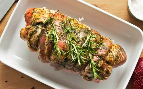 Rôti de porc Orloff, assaisonné et garni de romarin frais, prêt à être cuit dans un plat blanc.