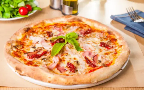 Pizza garnie de jambon, tomates séchées et champignons, parfaite pour une recette pâte à pizza rapide. Une salade fraîche est servie à côté.