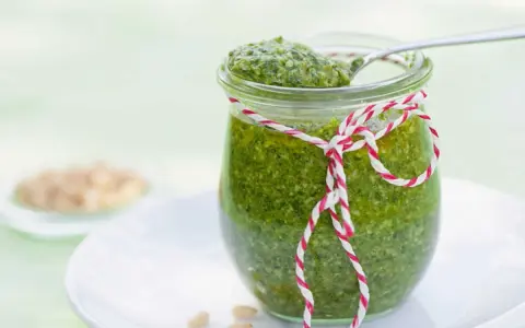 Un pot de pesto vert vif, décoré d'une ficelle rouge et blanche, prêt pour une recette pesto traditionnel. Quelques pignons de pin à côté.