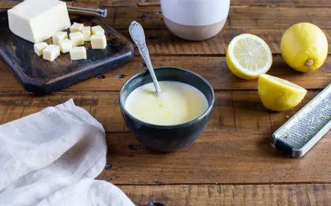 Un bol de sauce beurre citron, avec des citrons, du beurre coupé en dés et une râpe à zester sur une table en bois.