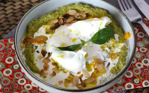 Un bol de purée verte, peut-être une sauce petit pois, garni d'un œuf poché, de crème, d'amandes effilées et d'une feuille de basilic.
