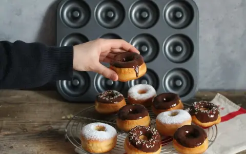 Une main tient un donut glacé au chocolat, prêt à être dégusté. Une belle collection de donuts au four, certains saupoudrés de sucre glace, d'autres de glaçage au chocolat et de vermicelles colorés, attendent sur une grille.