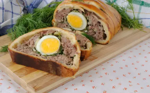 Une tranche de pâté de Pâques, garnie d'un œuf dur, est présentée sur une planche en bois, avec de l'aneth frais.
