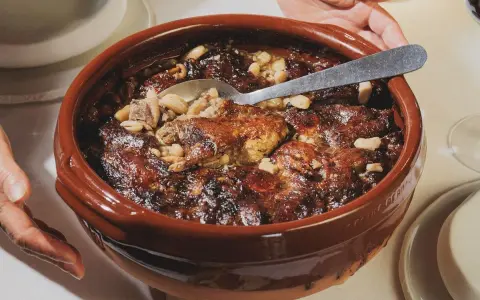 Un plat en terre cuite débordant de haricots blancs, de morceaux de viande dorée et de saucisses, prêt à être dégusté. La recette du vrai cassoulet traditionnel, un délice réconfortant.