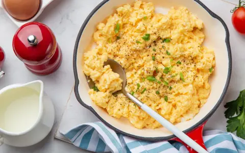 Un plat de oeufs brouillés, parsemé de ciboule et de poivre, servi dans une poêle rouge. Un moulin à poivre rouge et une cruche de lait sont à proximité.