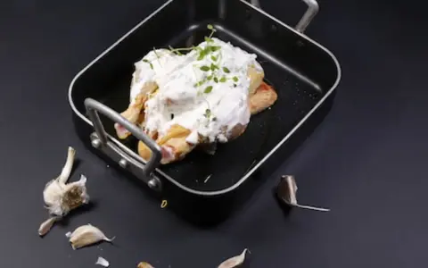 Un poulet entier est nappé de fromage blanc crémeux et garni d'herbes fraîches dans un plat noir. Des gousses d'ail sont disposées autour.