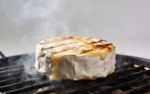 Un camembert au barbecue, fondant et fumant, cuit sur une grille. La croûte blanche se craquelle, révélant un cœur doré et coulant.