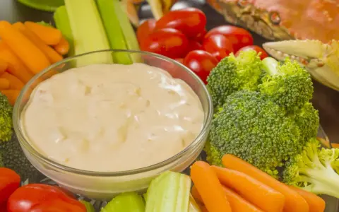 Un bol de sauce crudités crème fraîche entouré de carottes, céleri, tomates cerises et brocolis. Un crabe cuit est visible en arrière-plan.