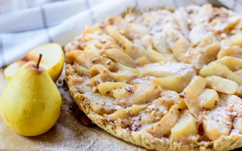 Délicieuse tarte aux poires, saupoudrée de sucre glace, avec des amandes et des poires fraîches à côté.