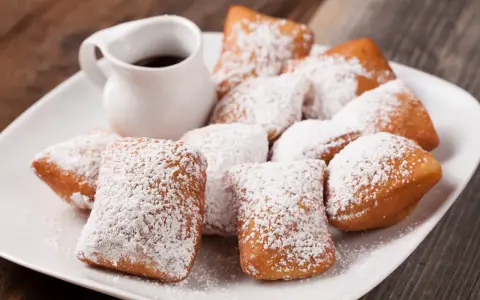 Une assiette de beignets saupoudrés de sucre glace, parfaits pour une recette pâte à beignets salés. Un petit pichet de sauce est à côté.