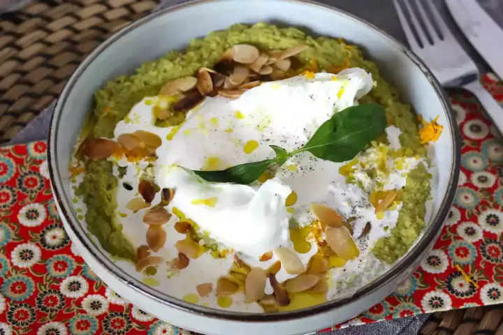 Un bol de purée verte, peut-être une sauce petit pois, garni d'un œuf poché, de crème, d'amandes effilées et d'une feuille de basilic.