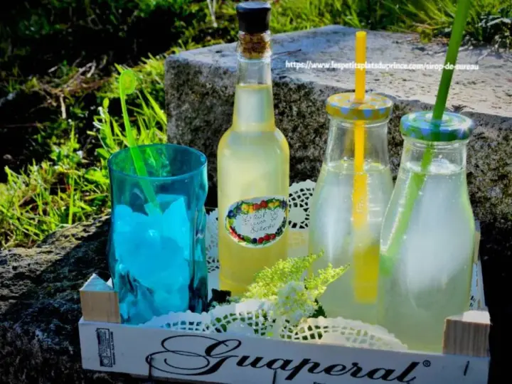 Bouteille de sirop de sureau et deux boissons rafraîchissantes avec des pailles, sur un plateau en bois, en extérieur.