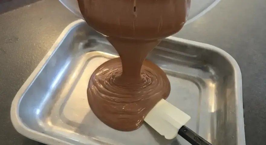 Comment faire une ganache : du chocolat fondu coule d'un bol dans un plat, guidé par une spatule.