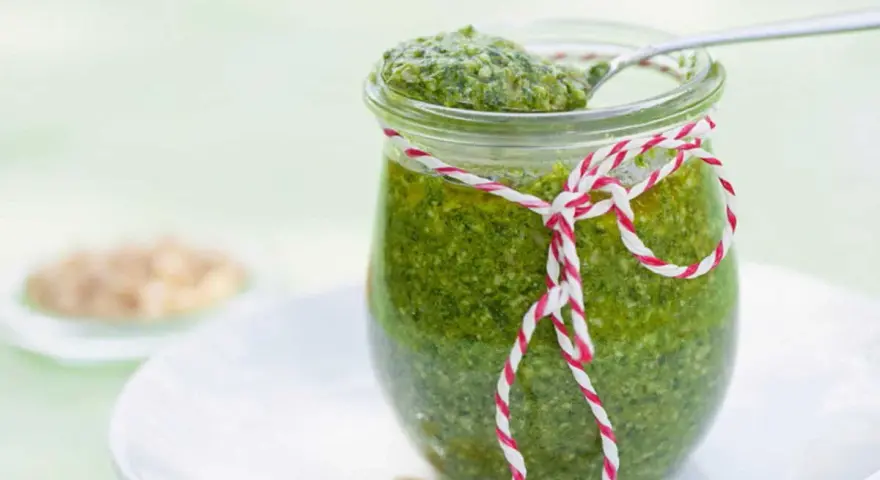 Un pot de pesto vert vif, décoré d'une ficelle rouge et blanche, prêt pour une recette pesto traditionnel. Quelques pignons de pin à côté.