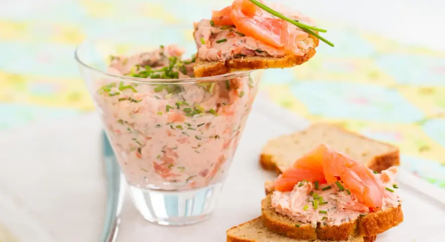 Rillettes de saumon crémeuses dans un verre, garnies de fines herbes et servies sur des toasts avec des tranches de saumon fumé.