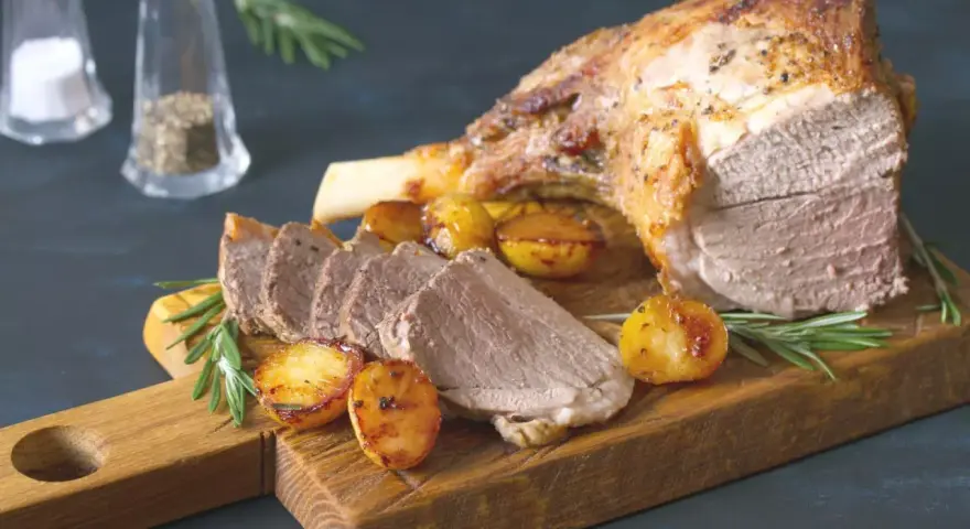 Un gigot de 7 heures, tendre et juteux, servi avec des pommes de terre rôties et du romarin sur une planche en bois.