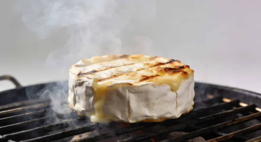 Un camembert au barbecue, fondant et fumant, cuit sur une grille. La croûte blanche se craquelle, révélant un cœur doré et coulant.