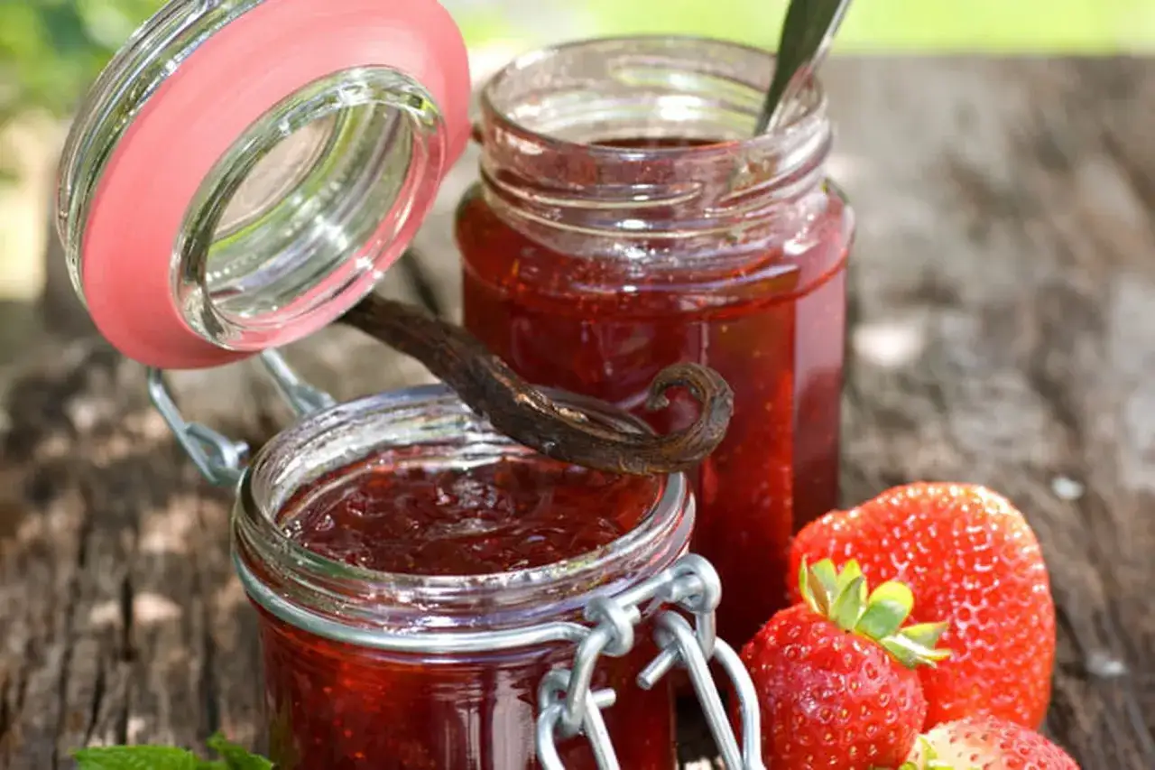 Deux pots de confiture de fraise maison, une gousse de vanille et des fraises fraîches sur une table en bois.
