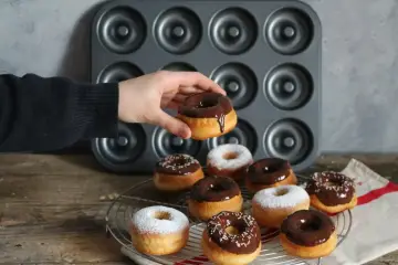 Une main tient un donut glacé au chocolat, prêt à être dégusté. Une belle collection de donuts au four, certains saupoudrés de sucre glace, d'autres de glaçage au chocolat et de vermicelles colorés, attendent sur une grille.